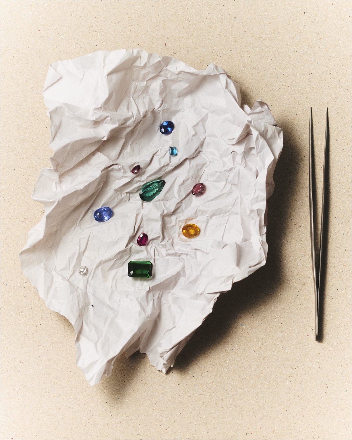 Loose coloured gemstones scattered across crumpled paper beside a pair of tweezers, selected during sourcing